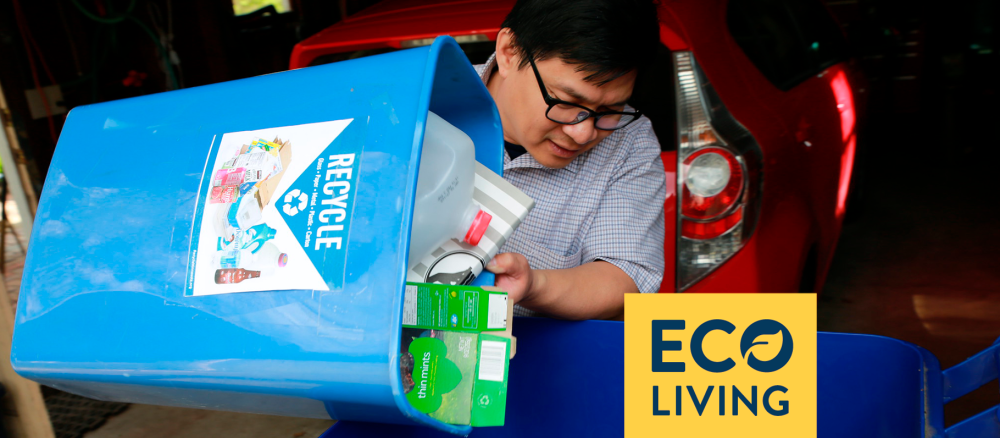 A man pours recycling, including a plastic jug and cardboard boxes, from a small blue bin into a larger one