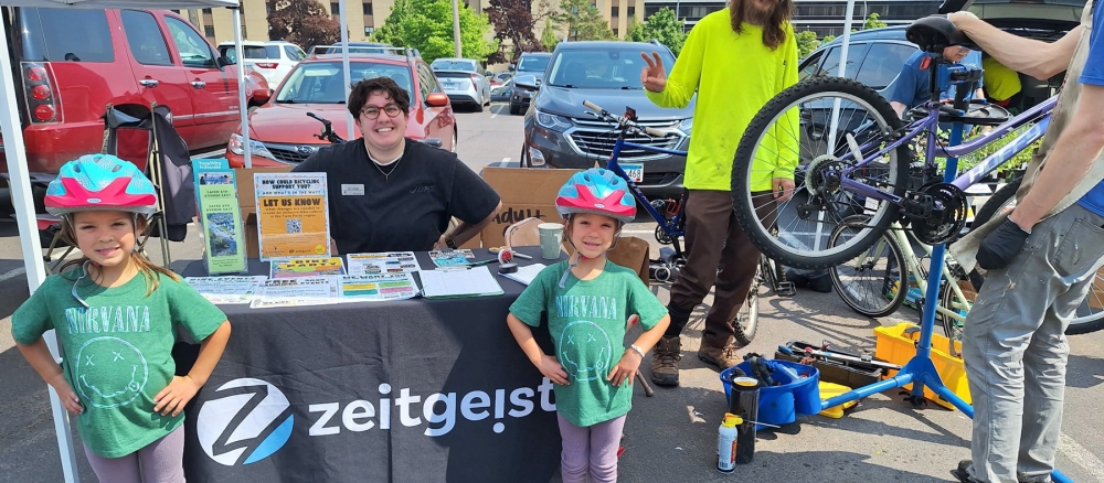 Two young girls wearing bike helmets stand in front of a table with promotional material. 