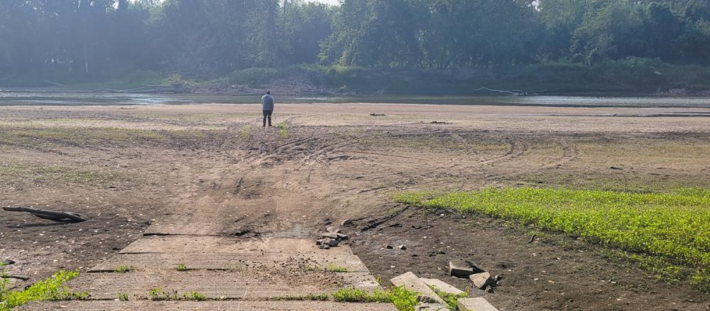 A boat ramp leads to a dry river bed. A man stands on the dry ground looking at the river in the distance.