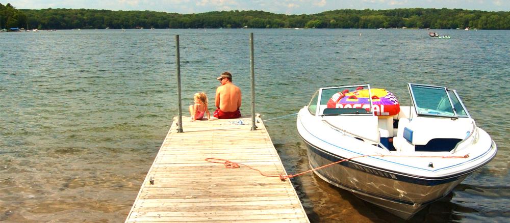 Father and daughter sitting on end of dock looking at lake. A white boat is tied to the side of the dock.
