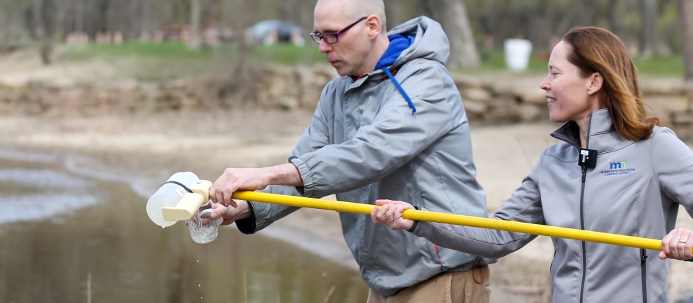 A man and woman next to a river pouring a water sample from a plastic container at the end of a long yellow pole into a glass jar.