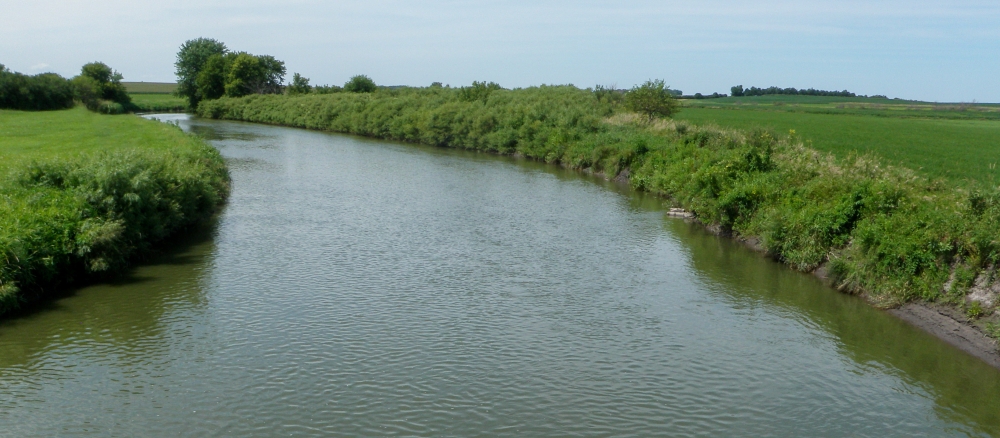 Wide, curving stream running through flat grasslands and fields. 