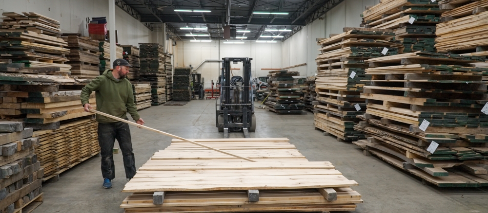 A man adds a board to a stack on a pallet in a warehouse. 