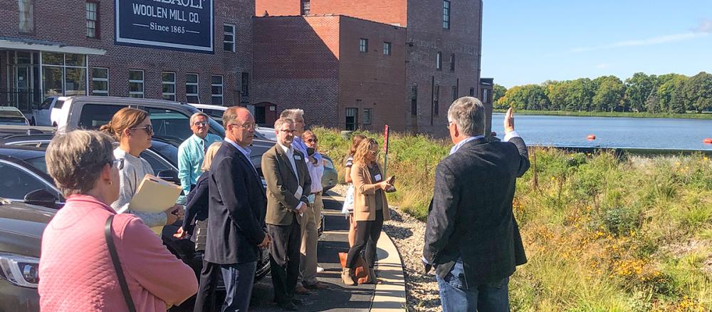 A man speaking to a group of people outside a brick building. The man is gesturing toward a rain garden and river in the distance.