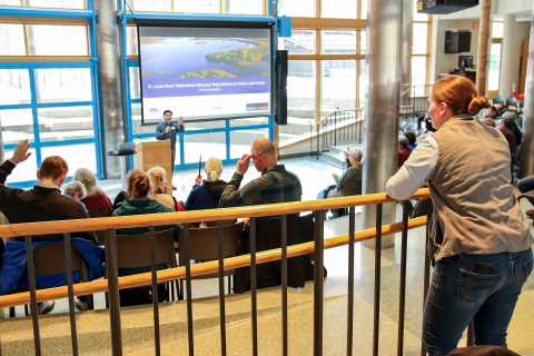 A woman standing at the back of an audience watching a man give a presentation.