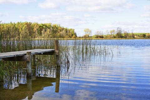 Old dock extends into small lake that is reflecting blue sky and clouds.