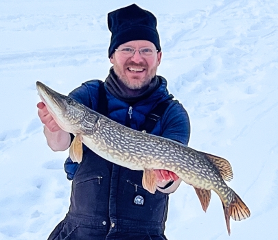 A smiling man standing on frozen lake holds up a large Northern Pike