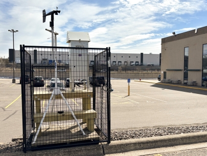 An air monitoring unit consisting of a white metal box and black propeller on a white tripod in a large parking lot behind a building.