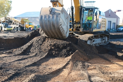 An excavator in an urban environment moving dirt into a pile to expose soil below.