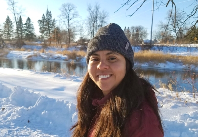Smiling woman in a coat and stocking hat stands in front of a river in winter.