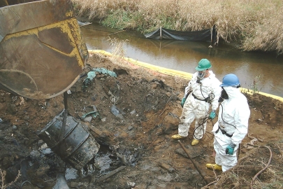 Men in hazardous waste suits observe an old leaking 50 gallon drum being pulled from the ground.