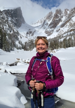 A smiling woman in a magenta jacket stands in front of a snow-covered mountain pass on a sunny day.