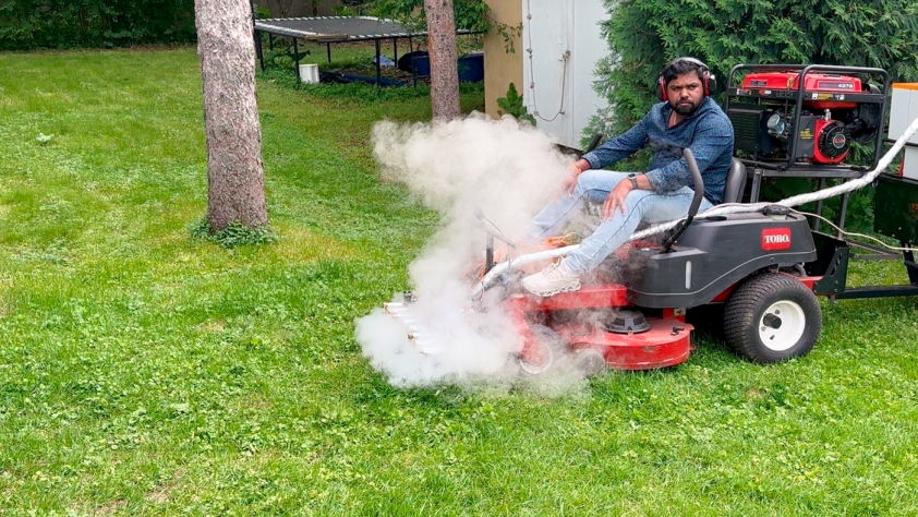 A man on a lawn mower with steam coming out of the front from special equipment attached to the mower.