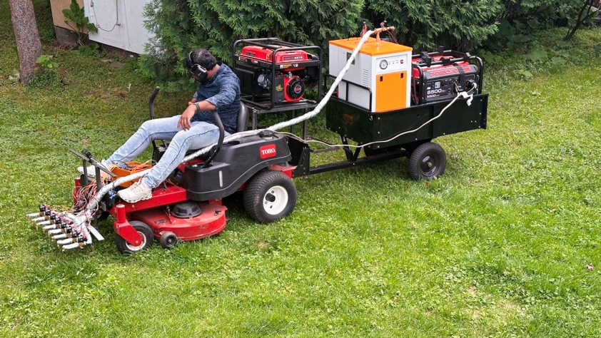 A man on a lawn mower with steam coming out of the front from special equipment attached to the mower.