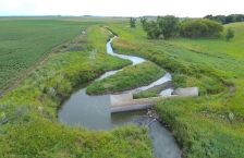 A small stream next to a farm field flows around one side of a concrete dam