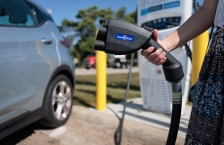 Person holding an EV charging connector. A car and EV charging station are in the background.