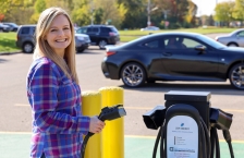 A smiling woman standing next to a electric vehicle charging station, holding the charging cord