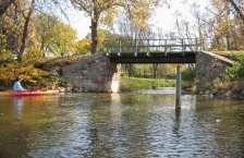 A person in a red and orange kayak floats down a calm stream, about to pass under a small foot bridge. The leaves on the surrounding trees are yellow and green.