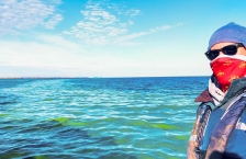 Man wearing winter clothes and sunglasses stands in front of large lake with a wide streak of bright green algae that runs to the horizon.