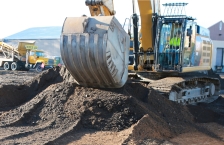 An excavator in an urban environment moving dirt into a pile to expose soil below.