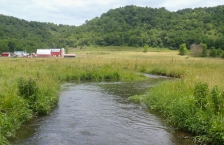Stream running through grassy field with red barn and farm buildings in background.