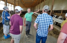 A man speaking to a group of people in a dairy barn with cows in the background.  