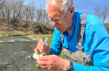 An older man in a blue shirt standing by a stream holding a tube of water.
