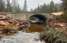 A cement bridge over a brown stream with a large culvert through it.