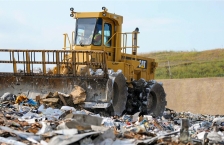 A yellow landfill compactor drives over trash in a landfill.