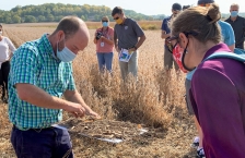 A man in a blue and green plaid shirt holds a white board with a pile of dried vegetation to explain the importance of crop residue to a group of people standing in a farm field.