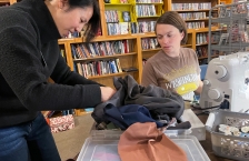 A woman looks through a stack of fabric as a woman sitting at a sewing machine watches.