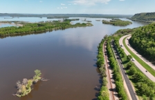 Aerial view of a large brown river with highways and railroads tracks running along the banks on the right side.