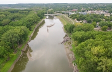 Aerial view of a brown river with tree-lined banks, a town on the right side, and a bridge in the distance.