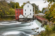 Old flour mill building next to dam with water flowing over it.