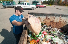 A man emptying a bag of cans and bottles into a recycling dumpster.