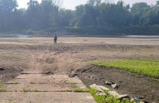 A boat ramp leads to a dry river bed. A man stands on the dry ground looking at the river in the distance.