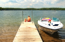 Father and daughter sitting on end of dock looking at lake. A white boat is tied to the side of the dock.