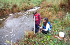 A woman and child stand next to a small stream. The child is looking into the top of a clear plastic tube.