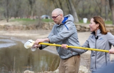 A man and woman next to a river pouring a water sample from a plastic container at the end of a long yellow pole into a glass jar.