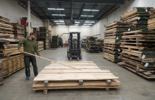 A man adds a board to a stack on a pallet in a warehouse. 