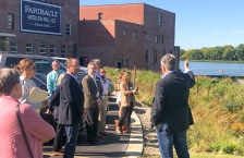 A man speaking to a group of people outside a brick building. The man is gesturing toward a rain garden and river in the distance.