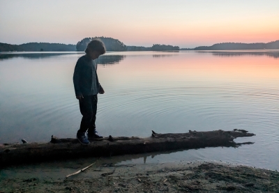 A child standing on a log on a lakeshore at sunset. 