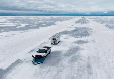 A white pickup drives across the ice pulling an ice house behind it. 