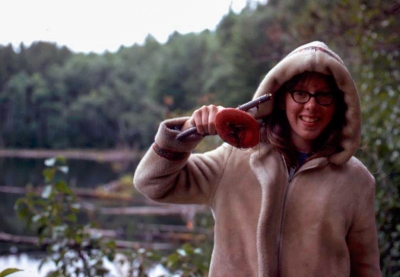 A young woman in a hooded coat stands in front of water holding up a stick and plastic disk. 