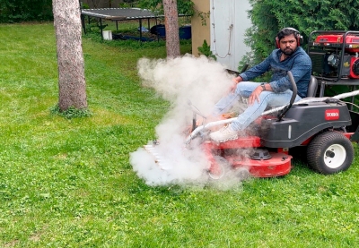 A man on a lawn mower with steam coming out of the front from special equipment attached to the mower. 