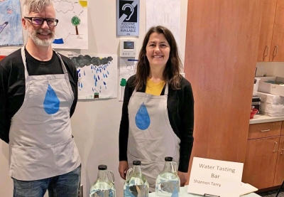 A man and a woman stand behind a table with 3 bottles of water that are used for taste testing samples from different areas of Otter Tail County.