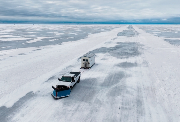 A white pickup drives across the ice pulling an ice house behind it. 