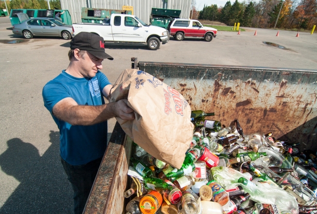 A man emptying a bag of cans and bottles into a recycling dumpster.
