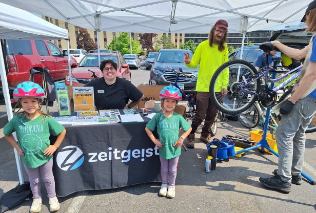 Two young girls wearing bike helmets stand in front of a table with promotional material. 