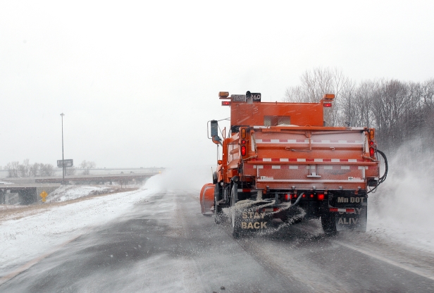 Rear view of an orange snow plow dropping salt on a snowy road.